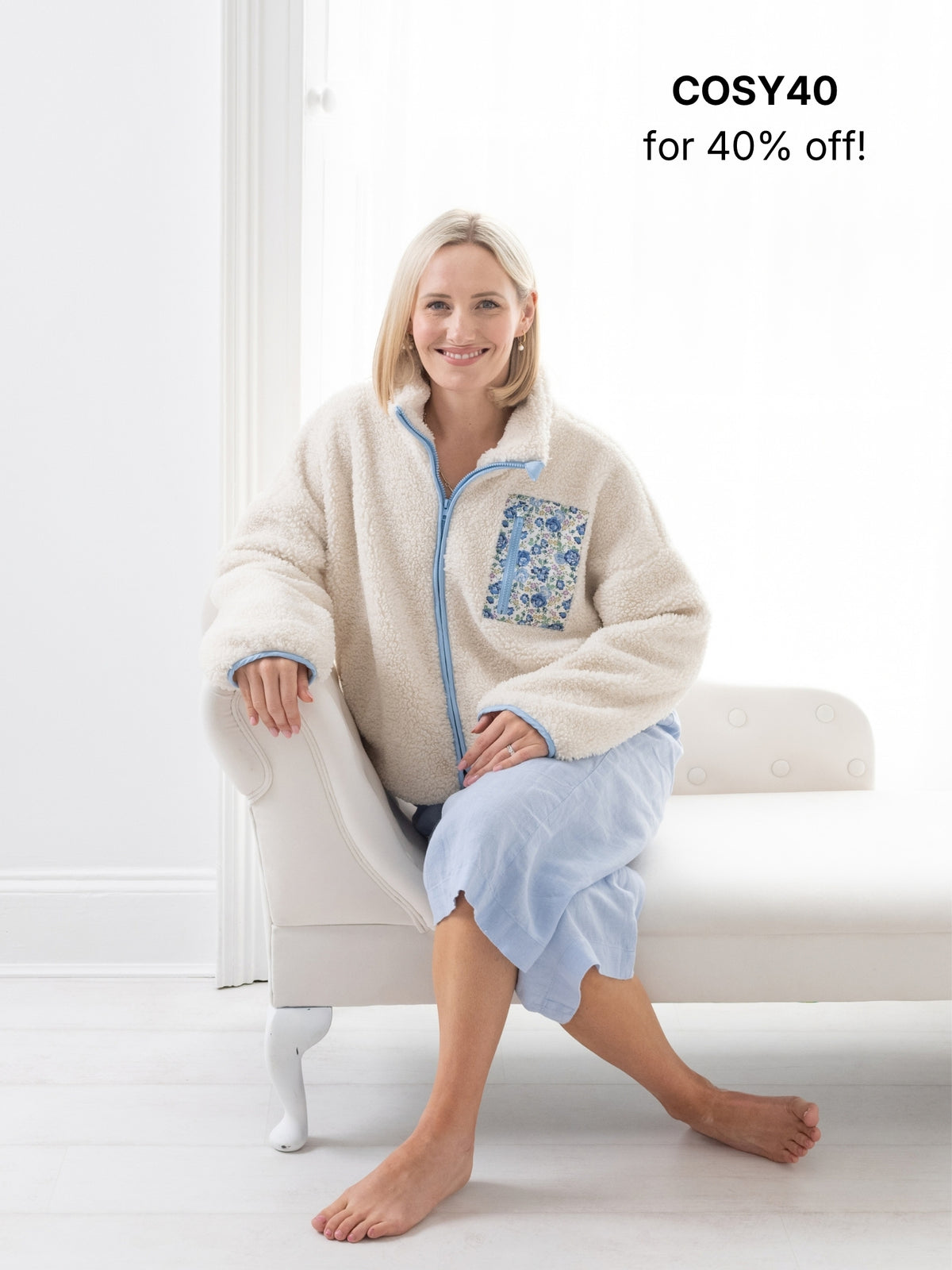 A woman sits barefoot on a white chaise lounge in a bright room, wearing a cream sherpa zip-up fleece jacket with a blue Liberty Print floral pocket and light blue culottes. Text in the top right reads 'COSY40 for 40% off!'.