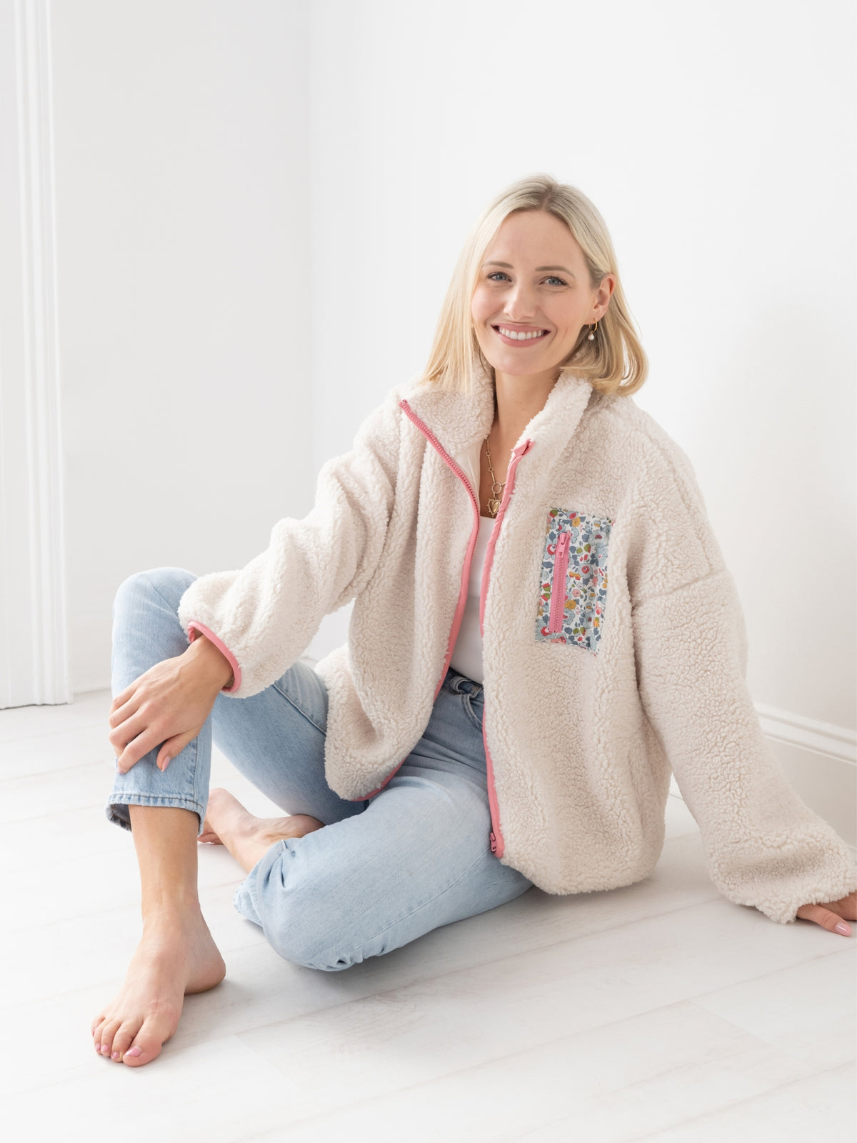 A smiling blonde woman sits barefoot on a white floor in a bright room, wearing a cream sherpa borg fleece zip-up jacket with pink trim and a Liberty Print floral patch pocket, paired with light blue jeans.