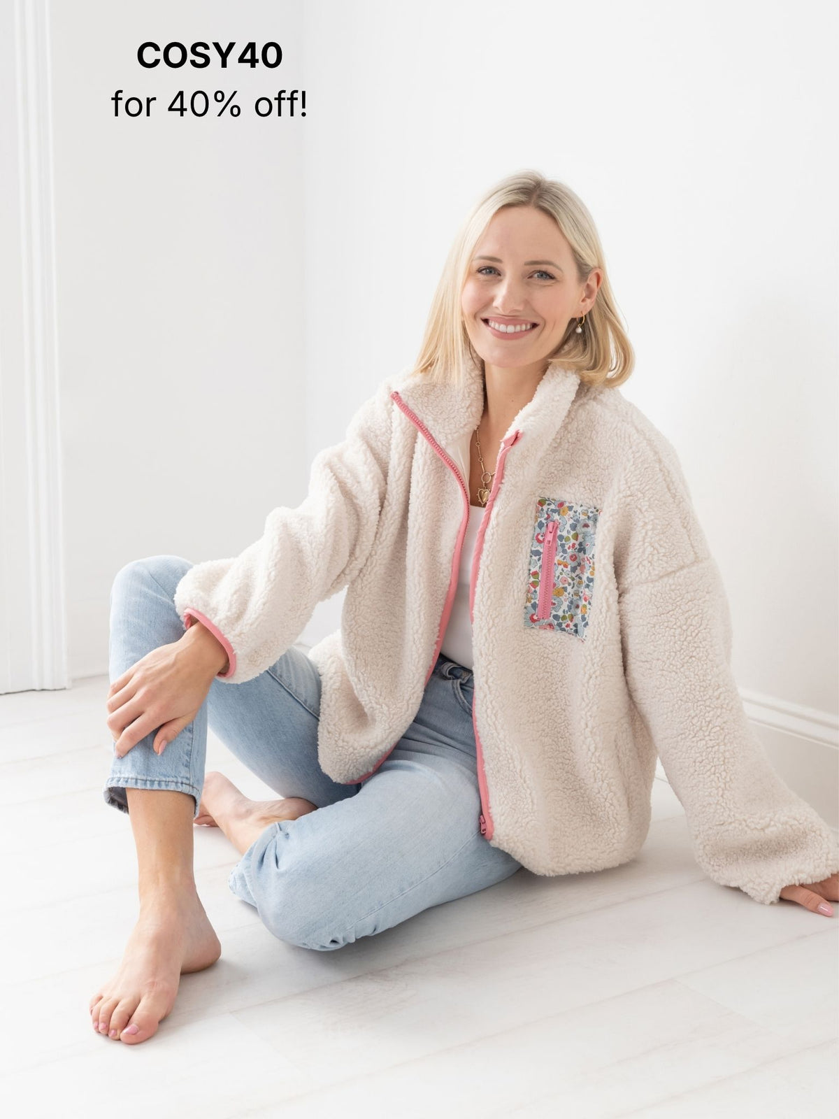 A woman sits barefoot on a white floor wearing a cream fleece sherpa zip-up jacket with pink trim and a floral patch pocket, paired with light blue jeans. Text in the top left reads 'COSY40 for 40% off!'.